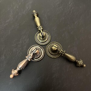 May include: Three antique brass-toned metal drawer pulls arranged in a triangular formation. Each pull features a circular backplate with a beaded edge and a hinged, teardrop-shaped handle with decorative filigree detailing at the base and tip.