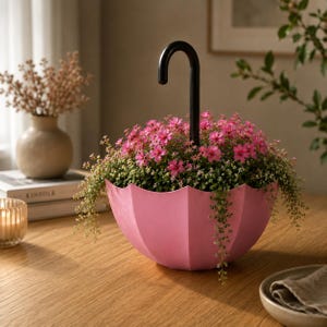 May include: A pink umbrella-shaped planter containing a dense arrangement of small pink flowers and green foliage. The planter features a curved black handle extending upwards from the centre, mimicking the appearance of an inverted umbrella.