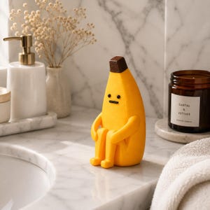 Puede incluir: Una figura de plátano amarillo con la punta del tallo marrón, ojos de puntos negros y una línea negra recta como boca. El plátano está en posición sentada con los brazos alrededor de las rodillas, con un acabado mate y liso.