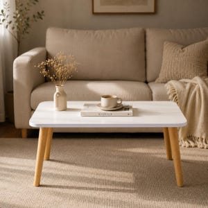 Peut inclure: Une table basse rectangulaire blanche avec quatre pieds en bois clair. Sur la table se trouvent un petit vase en céramique beige contenant des fleurs séchées, un livre blanc intitulé KINFOLK et une tasse à café blanche sur sa soucoupe.