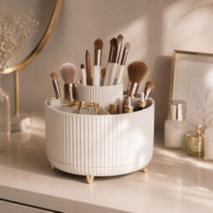 Könnte beinhalten: Ein weißer, zylindrischer Make-up-Organizer mit gerippter Oberfläche, einem erhöhten Mittelteil und mehreren Außenfächern. Er steht auf kleinen goldfarbenen Füßen und ist mit verschiedenen Make-up-Pinseln, Lippenstiften und kleinen Kosmetikfläschchen gefüllt.