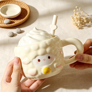 Puede incluir: Una taza de cerámica color crema con forma de oveja esponjosa, superficie texturizada, asa y una tapa que simula nata montada. La taza incluye una pajita a juego con un pequeño detalle de corazón y presenta una cara de oveja con mejillas rosas.