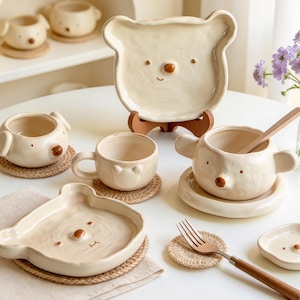 Peut inclure: Une collection de vaisselle en céramique couleur crème avec des motifs en forme d'ours, comprenant des assiettes, des bols et des tasses avec de petits nez et oreilles marron. L'ensemble inclut plusieurs pièces assorties sur des dessous de verre.