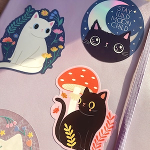 May include: Three cat stickers with different designs. One sticker features a white cat with blue eyes surrounded by flowers. Another sticker features a black cat with blue eyes and a crescent moon with the text "Stay Wild Moon Child". The third sticker features a black cat sitting under a red mushroom with white spots.