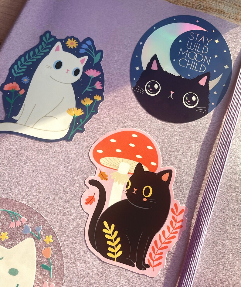 May include: Three cat stickers with different designs. One sticker features a white cat with blue eyes surrounded by flowers. Another sticker features a black cat with blue eyes and a crescent moon with the text "Stay Wild Moon Child". The third sticker features a black cat sitting under a red mushroom with white spots.