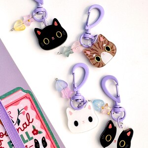 May include: Four cat-themed keychains with purple plastic lobster clasps and small star and heart-shaped beads. The charms feature a black cat, a brown tabby cat, a white cat, and a black and white tuxedo cat, all with large eyes and whiskers.