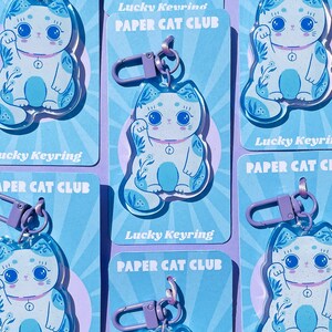 Pu&ograve; includere: Un set di sei portachiavi a forma di gatto fortunato blu e bianco con motivi floreali. Ogni portachiavi ha un anello di metallo viola e un ciondolo di plastica trasparente. I ciondoli raffigurano un gatto bianco con occhi blu e naso rosa, che alza la zampa destra. I ciondoli sono stampati con il testo "Lucky Keyring Paper Cat Club".