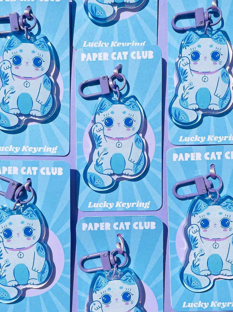 Pu&ograve; includere: Un set di sei portachiavi a forma di gatto fortunato blu e bianco con motivi floreali. Ogni portachiavi ha un anello di metallo viola e un ciondolo di plastica trasparente. I ciondoli raffigurano un gatto bianco con occhi blu e naso rosa, che alza la zampa destra. I ciondoli sono stampati con il testo "Lucky Keyring Paper Cat Club".