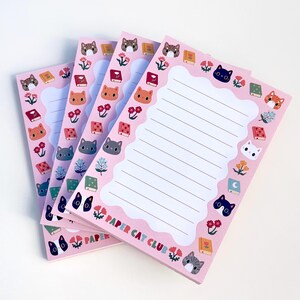 May include: A stack of rectangular pink notepads with a scalloped white lined writing area in the center. The border features illustrations of various cat faces, colorful flowers, and small books. The text PAPER CAT CLUB is printed at the bottom.