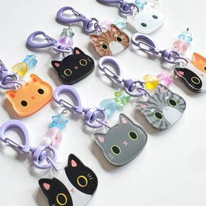 May include: A collection of acrylic cat-shaped keychains, each featuring a unique feline face design in colors including black, orange, gray, and tabby. Each keychain includes a purple plastic lobster clasp and small star and heart-shaped beads.