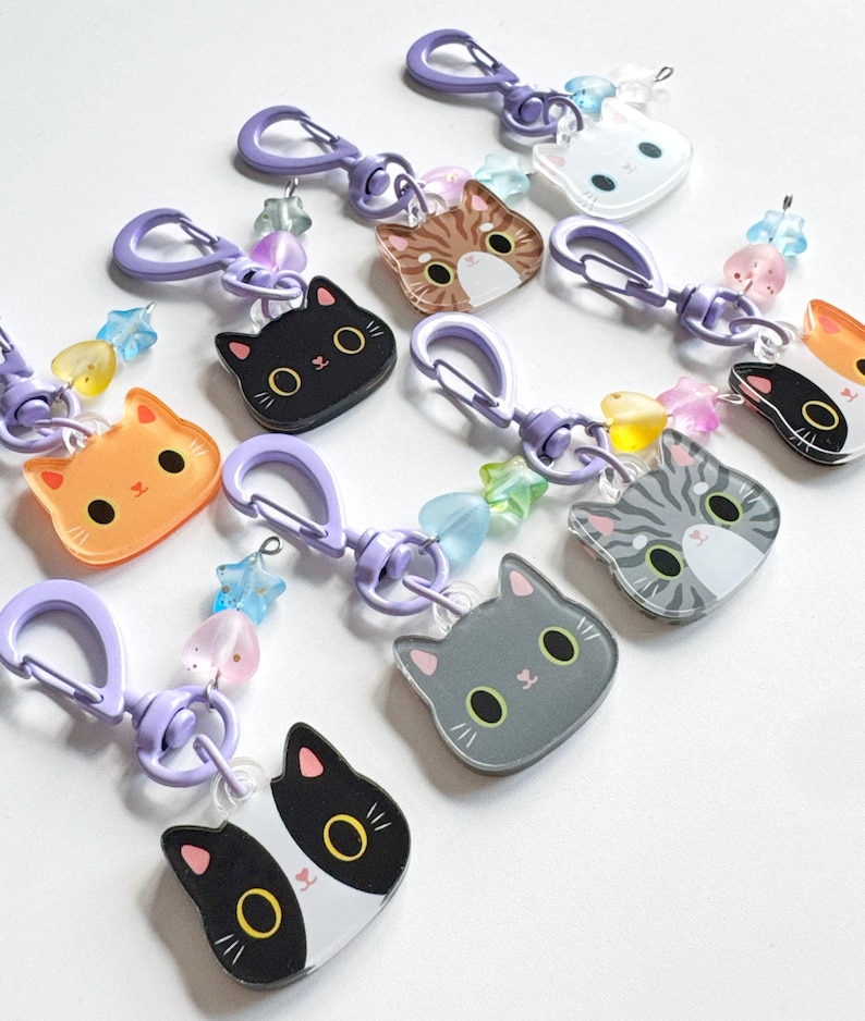 May include: A collection of acrylic cat-shaped keychains, each featuring a unique feline face design in colors including black, orange, gray, and tabby. Each keychain includes a purple plastic lobster clasp and small star and heart-shaped beads.
