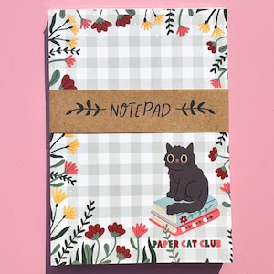 May include: A notepad with a grey and white gingham pattern, floral border, and a brown band that reads "NOTEPAD." The notepad features a black cat sitting on a stack of books, with the text "PAPER CAT CLUB."