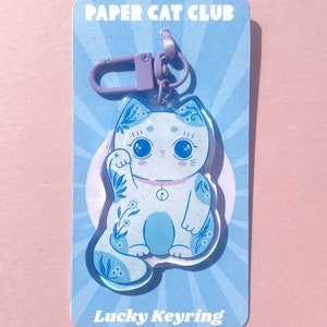 Pu&ograve; includere: Un portachiavi a forma di gatto fortunato blu e bianco con un design floreale. Il portachiavi &egrave; su una carta blu e bianca con il testo "Paper Cat Club" e "Lucky Keyring".