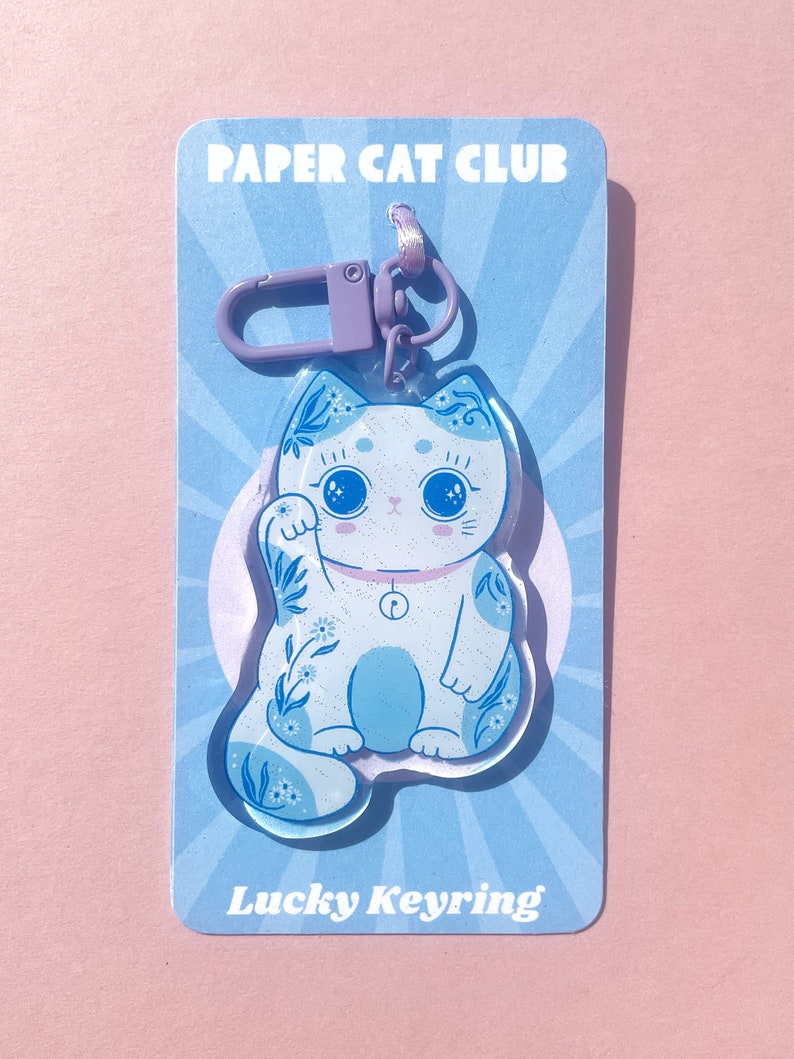 Pu&ograve; includere: Un portachiavi a forma di gatto fortunato blu e bianco con un design floreale. Il portachiavi &egrave; su una carta blu e bianca con il testo "Paper Cat Club" e "Lucky Keyring".