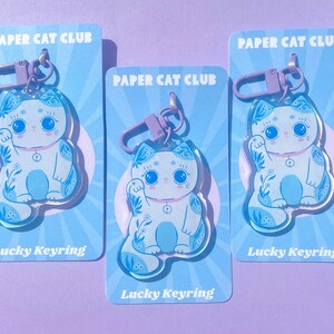 Pu&ograve; includere: Tre portachiavi a forma di gatto portafortuna blu e bianco con motivi floreali. Ogni portachiavi &egrave; attaccato a un portachiavi in metallo viola e ha il testo "PAPER CAT CLUB" e "Lucky Keyring" stampato sulla carta.