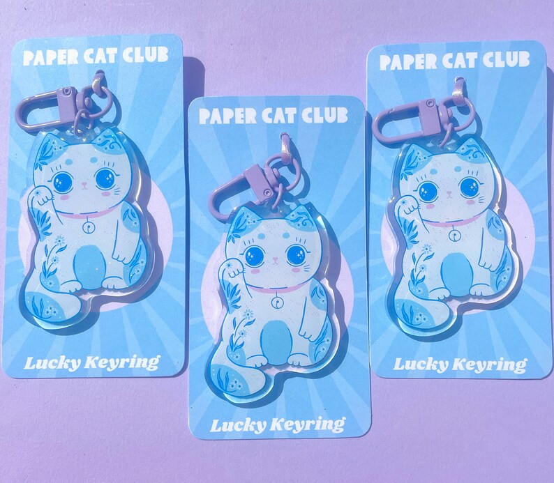 Pu&ograve; includere: Tre portachiavi a forma di gatto portafortuna blu e bianco con motivi floreali. Ogni portachiavi &egrave; attaccato a un portachiavi in metallo viola e ha il testo "PAPER CAT CLUB" e "Lucky Keyring" stampato sulla carta.