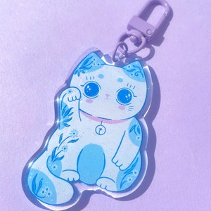 Pu&ograve; includere: Un portachiavi in metallo viola con un ciondolo a forma di gatto blu e bianco con glitter e un campanello al collo.