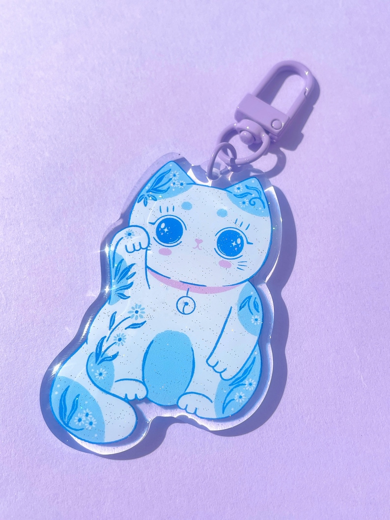 Pu&ograve; includere: Un portachiavi in metallo viola con un ciondolo a forma di gatto blu e bianco con glitter e un campanello al collo.