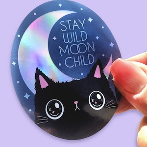 May include: A round sticker featuring a black cat with pink ears and large eyes. The cat is positioned in front of a crescent moon with a holographic finish. The text "STAY WILD MOON CHILD" is written in white against a blue background.