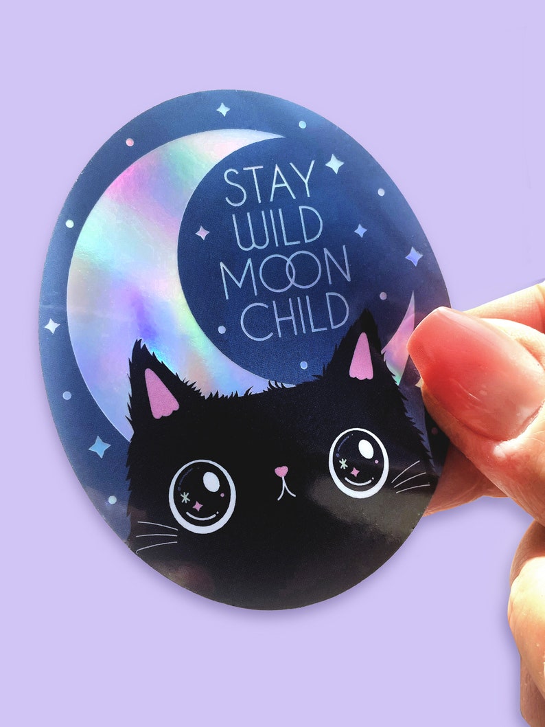 May include: A round sticker featuring a black cat with pink ears and large eyes. The cat is positioned in front of a crescent moon with a holographic finish. The text "STAY WILD MOON CHILD" is written in white against a blue background.