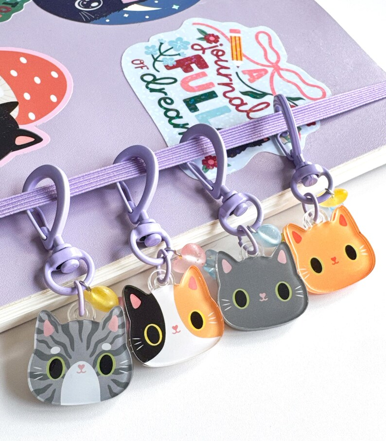 May include: Four acrylic cat head charms attached to light purple swivel lobster clasps. The charms feature a gray tabby, a calico, a solid gray cat, and an orange cat. Each charm includes a small colorful bead and hangs from a purple elastic band.