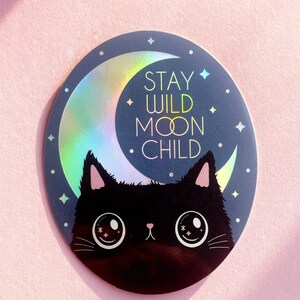 May include: A black cat with large eyes peeks out from behind a crescent moon with a holographic finish. The moon has the text "Stay Wild Moon Child" on it.