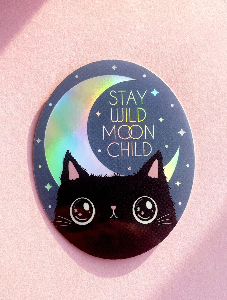May include: A black cat with large eyes peeks out from behind a crescent moon with a holographic finish. The moon has the text "Stay Wild Moon Child" on it.