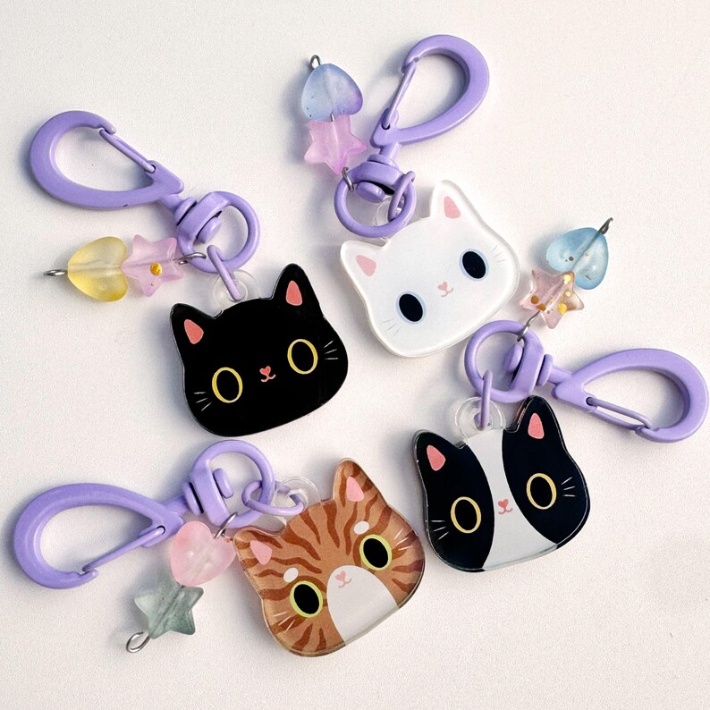 May include: Four cat-shaped acrylic keychains with purple plastic lobster clasps. Each keychain features a unique cat face design, including black, white, orange tabby, and tuxedo patterns, accented with small translucent heart and star beads.