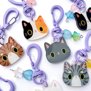 May include: A collection of acrylic cat-shaped keychains, each featuring a unique feline face design. The keychains are attached to light purple plastic lobster clasps and accented with small, colorful star and heart-shaped beads.