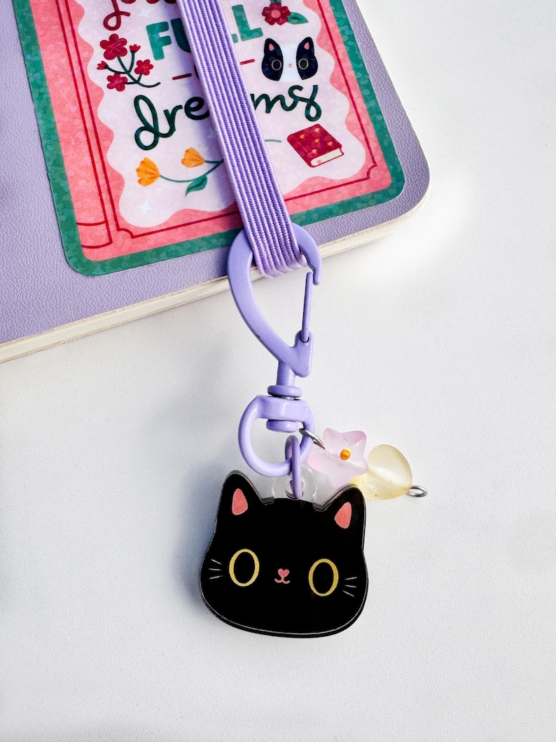 May include: A lavender elastic strap with a matching plastic lobster clasp attached to a black cat-shaped acrylic charm. The charm features large yellow eyes, pink inner ears, and a small pink nose. Two small translucent flower and heart beads hang nearby.