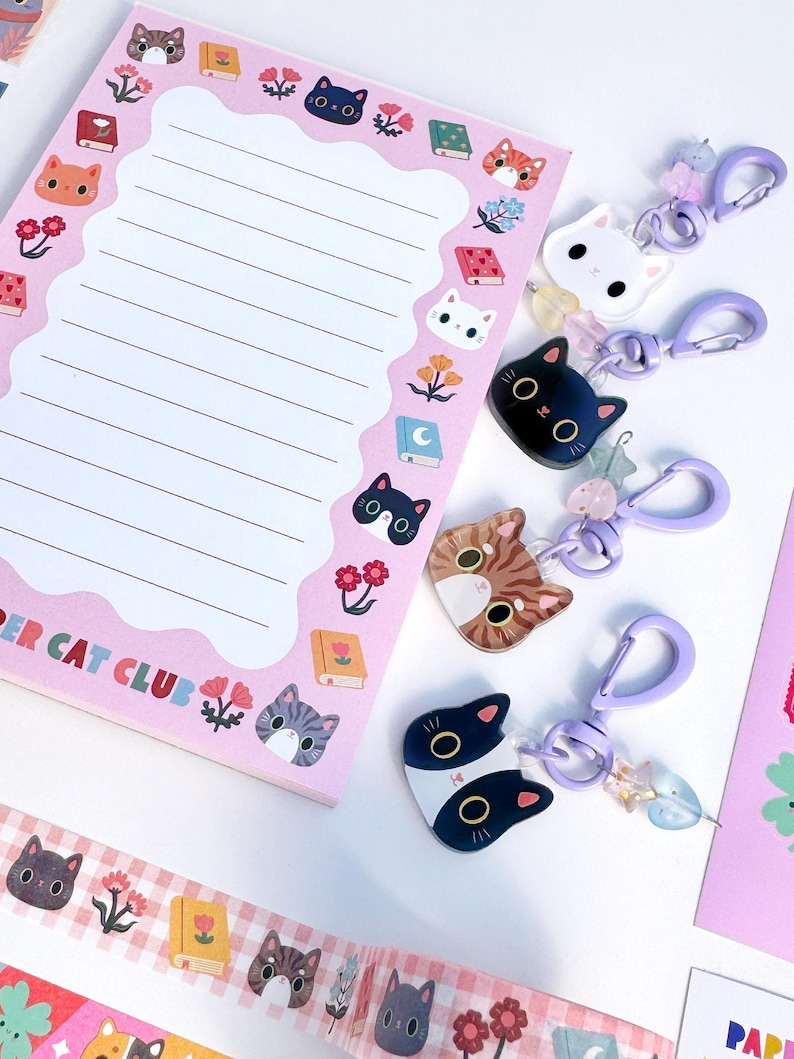 May include: A pink notepad with lined paper and a border featuring illustrations of cats, books, and flowers, with the text PAPER CAT CLUB. Four cat-shaped acrylic keychains with purple clasps and small star-shaped beads are arranged to the right.