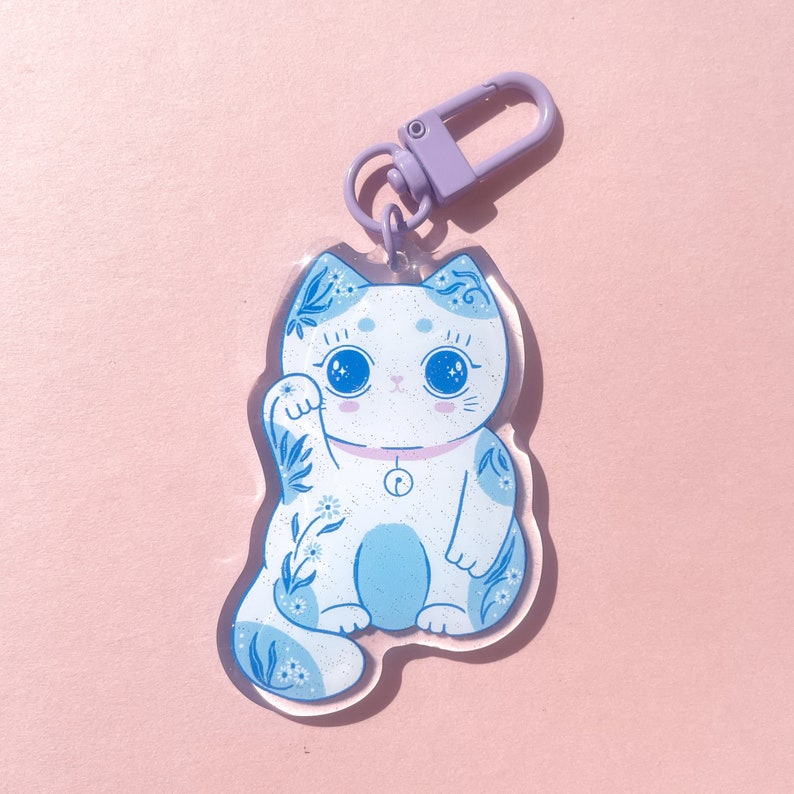 Pu&ograve; includere: Un ciondolo a forma di gatto fortunato blu e bianco con un portachiavi viola. Il gatto alza la zampa e ha un disegno floreale sul corpo.