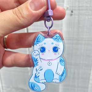 Pu&ograve; includere: Un ciondolo Maneki-Neko, o gatto portafortuna, blu e bianco con un colletto rosa e un campanello. Il ciondolo &egrave; attaccato a un portachiavi viola.