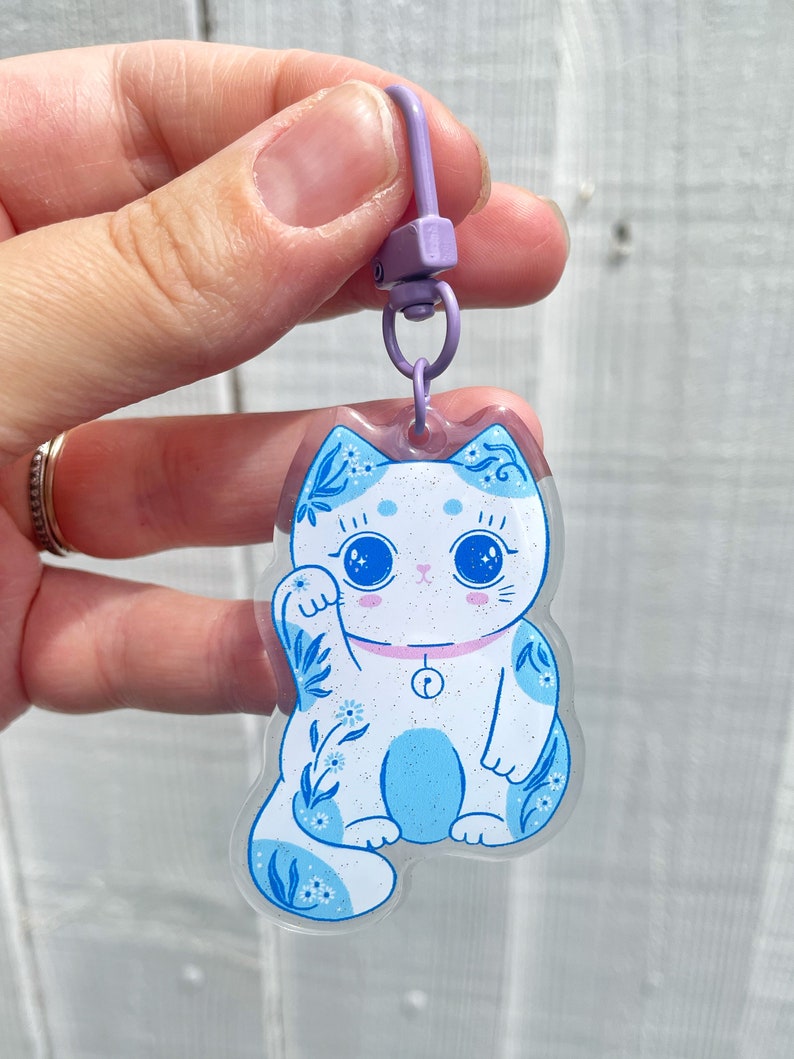 Pu&ograve; includere: Un ciondolo Maneki-Neko, o gatto portafortuna, blu e bianco con un colletto rosa e un campanello. Il ciondolo &egrave; attaccato a un portachiavi viola.