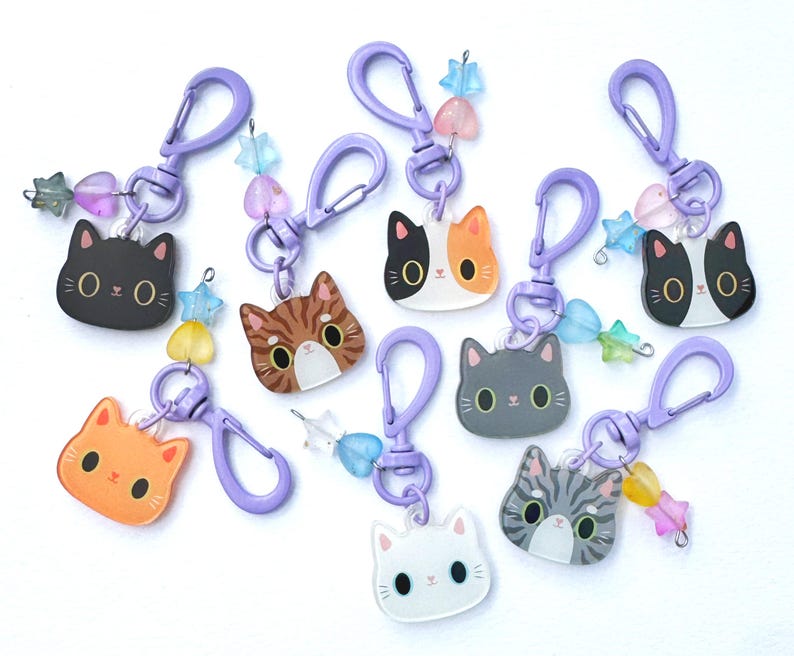 May include: A collection of eight cat-themed keychains featuring acrylic cat head charms in various colors and patterns, including black, orange, tabby, and calico. Each keychain has a purple plastic lobster clasp and small star and heart beads.