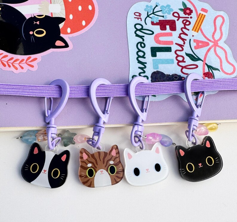 May include: Four cat-shaped acrylic charms attached to light purple plastic lobster clasps. The charms feature a black and white cat, a brown tabby cat, a white cat, and a black cat. Each charm includes small pastel-colored star and bead accents.