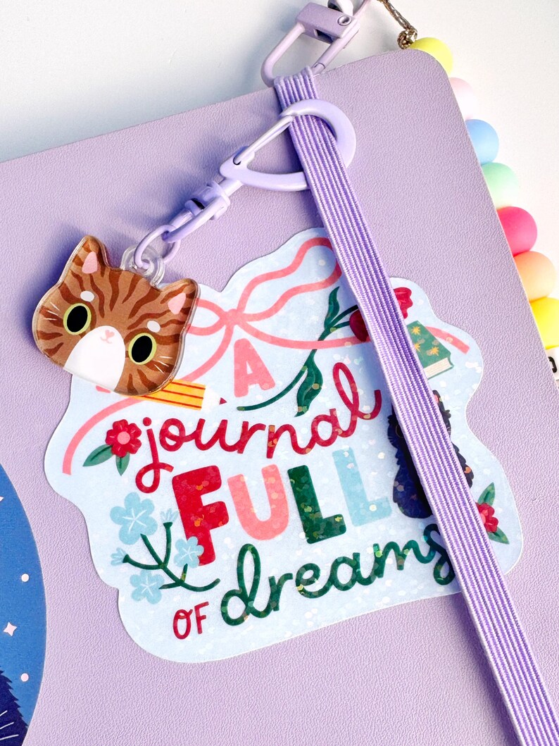 May include: A lavender journal with a light purple elastic band and a light purple plastic clip. Attached to the clip is an acrylic charm of a brown tabby cat head and a sticker featuring the text A journal FULL of dreams with floral art.