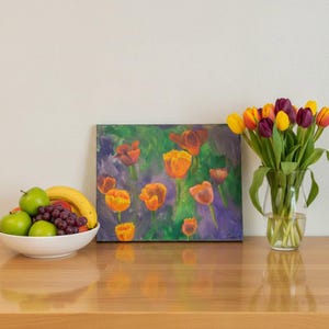 May include: A rectangular painting featuring orange tulips against a green and purple background, positioned next to a white bowl filled with green apples, a yellow banana, and purple grapes, and a clear glass vase containing yellow and purple tulips.