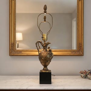 May include: A metallic gold-toned lamp base shaped like an ornate pitcher with intricate embossed scrollwork and a curved handle. The lamp features a brass harp assembly with a finial on top, all mounted on a solid black square base.