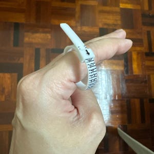 May include: A white plastic ring sizer tool featuring a buckle mechanism and a printed scale with black numerical markings ranging from 11 to 16, designed to measure finger circumference for determining accurate ring sizes.