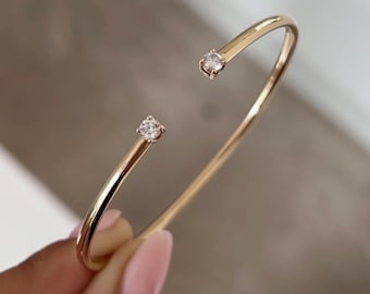 May include: A gold-toned open cuff bracelet featuring a smooth, polished metal band. The two ends of the bracelet are each finished with a single, small, clear, round-cut gemstone set in a prong setting, creating a minimalist and elegant design.