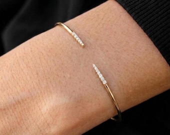 May include: A gold open cuff bracelet featuring two tapered ends, each set with a row of small, clear, round-cut stones. The thin, polished gold band curves around the wrist, leaving a gap between the two embellished, pointed terminals.