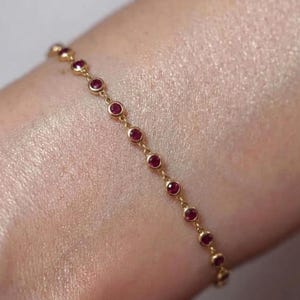 May include: A delicate gold chain bracelet featuring a series of small, round, bezel-set red gemstones spaced evenly along the length of the chain. The bracelet is constructed with thin gold links connecting each individual circular gemstone setting.