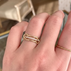 Op de afbeelding: Een gouden ring met twee parallelle horizontale banden verbonden door lusvormige uiteinden, met een centrale balk ingelegd met kleine heldere steentjes. Een tweede dunne gouden ring met een kralentextuur wordt aan de naastgelegen vinger gedragen.