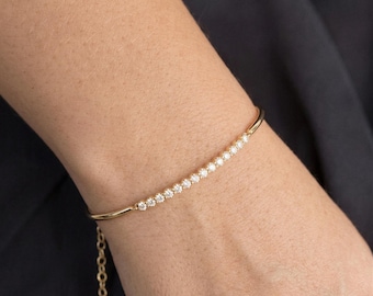 May include: A gold-toned bracelet featuring a curved bar set with a row of small, clear, round-cut stones. The bracelet includes a delicate chain extension with circular links, providing an adjustable fit for the wrist.