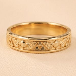 May include: A gold-toned band ring featuring a repeating engraved pattern of triangular Triforce symbols and hexagonal shapes. The metal surface has a polished finish on the edges and a textured, matte appearance within the central band design.