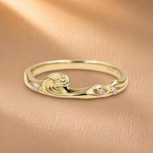 Op de afbeelding: Een gouden ring met een sculpturaal golfontwerp dat rond de band loopt. Het metalen oppervlak heeft een gepolijste afwerking en is geaccentueerd met drie kleine, ronde, heldere edelstenen die langs de vloeiende, organische rondingen zijn ingelegd.