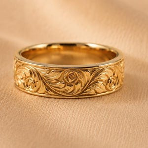 May include: A gold-toned band ring featuring an intricate, continuous engraved scrollwork pattern that wraps around the entire exterior surface. The metal has a polished finish that reflects light, highlighting the deep, carved floral-inspired details.