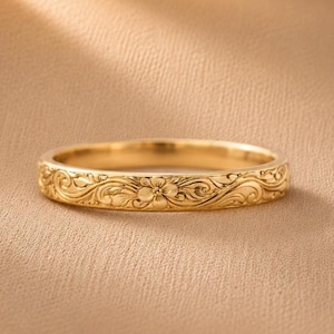 May include: A gold band ring featuring an intricate, embossed floral and scrollwork pattern that wraps around the entire exterior surface. The metal has a polished finish, highlighting the detailed engravings of small flowers and swirling vines.