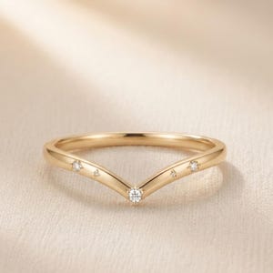 May include: A V-shaped gold ring featuring a polished band with five small, clear, round-cut stones set along the top curve. The stones are arranged with a larger centre stone at the point of the V, flanked by two smaller stones and two tiny star-shaped accents.